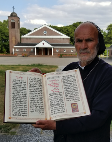 Dayroyo Elio Atto holding an open copy of his handwritten Gospel Lectionary in front of a church